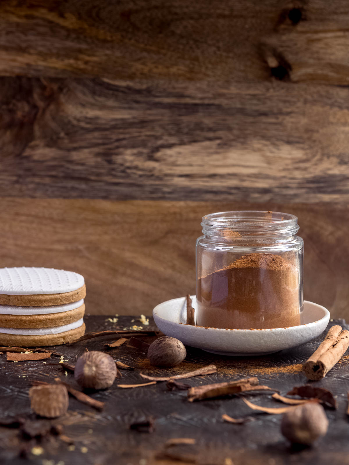 A small jar of homemade speculaas spice mix sits on a white plate. Nutmeg and cinnamon surround it and there's 3 speculaas sugar cookies covered in fondant stacked on the side