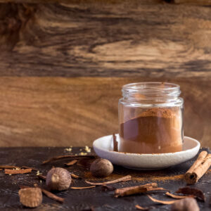 A small glass jar filled with homemade speculaas spice mix, sits on a white plate, surrounded with nutmegg, cinnamon quills and mixed spices