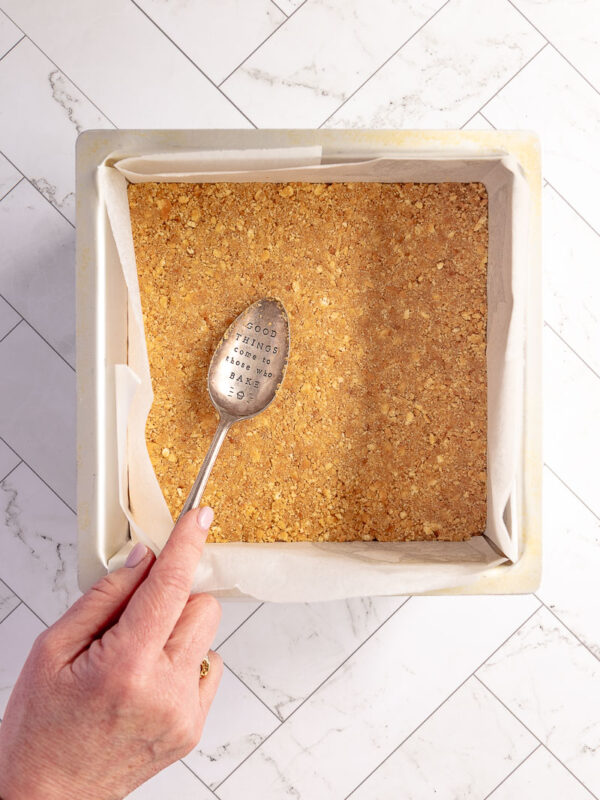 using a metal spoon to press the biscuit base into a square tin