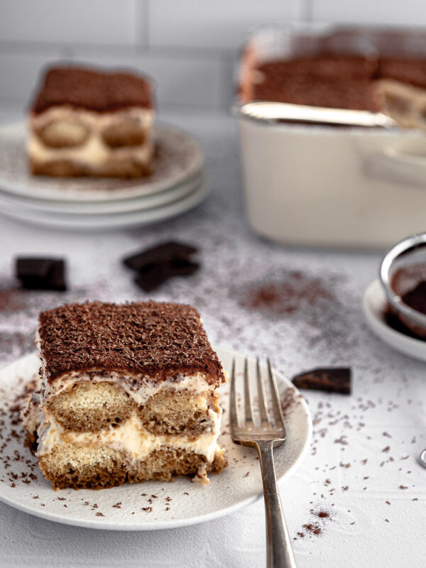 a slice of tiramisu on a white plate with a vintage fork beside it. A serving bowl of tiramisu is in the background, along with a stack of plates