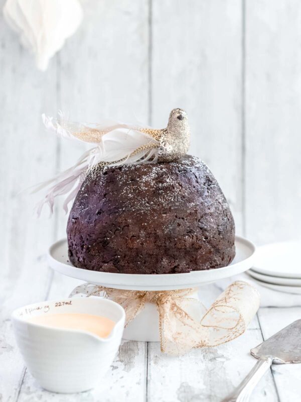 traditional christmas pudding dusted with icing sugar and sitting on a pedestal cake stand. A gold bird sits on top