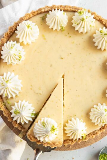A whole gluten-free Key Lime Pie with a wedge being served, topped with whipped cream and garnished with lime zest.