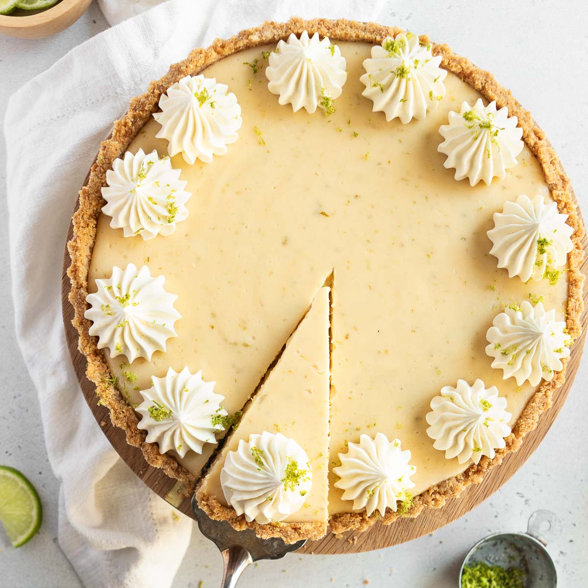 A whole gluten-free Key Lime Pie with a wedge being served, topped with whipped cream and garnished with lime zest.
