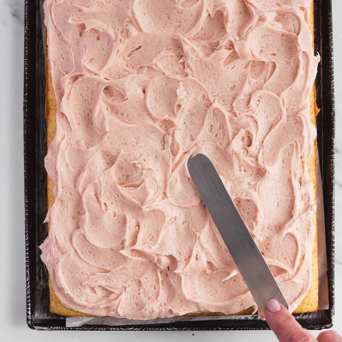 Fluffy vanilla sheet cake being frosted with a strawberry buttercream.