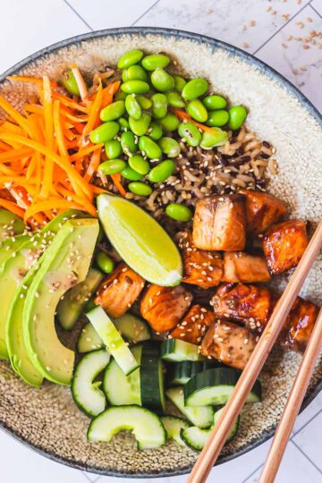 Sticky honey soy salmon served in a bowl, along with edamame, rice, avocado, carrot and cucumber.